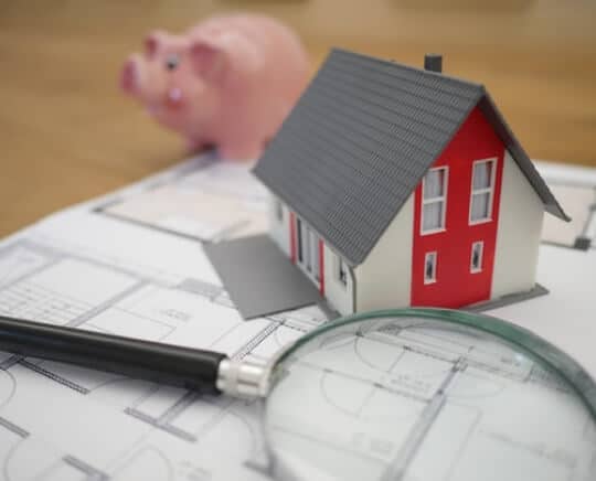 A piggy bank, magnifying glass, and small paper home on table with a blueprint